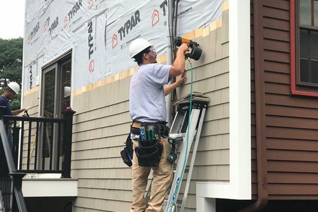 Construction worker in a hard hat and safety gear carefully measuring and installing tan house siding over home insulation.