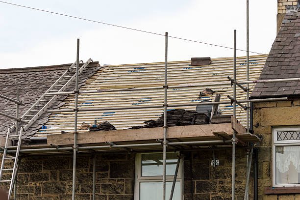 A man is re-roofing a house, working diligently on the roof under clear blue skies.