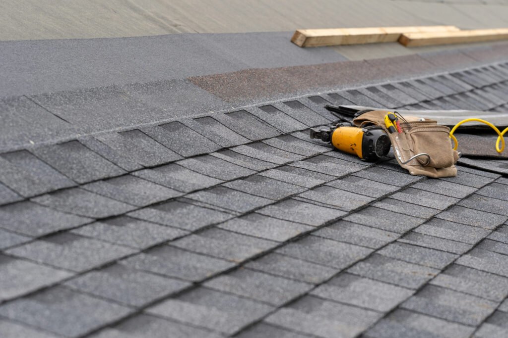 A man in a yellow vest installs asphalt shingles on a roof under clear blue skies.