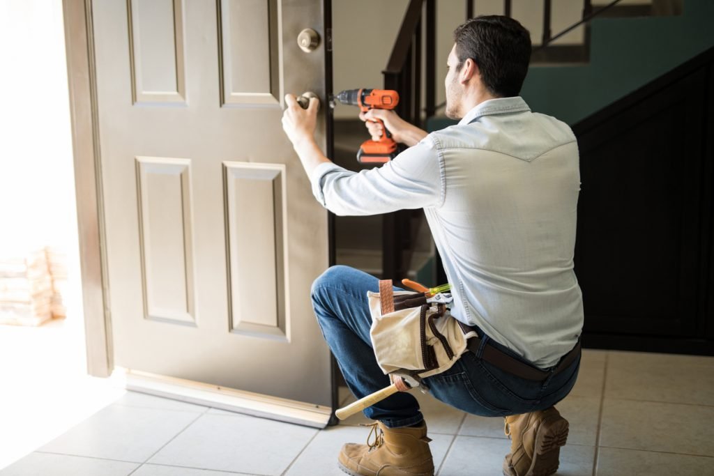 A man in a light blue shirt and work boots kneels to install a doorknob on a wooden front door using an orange power drill.