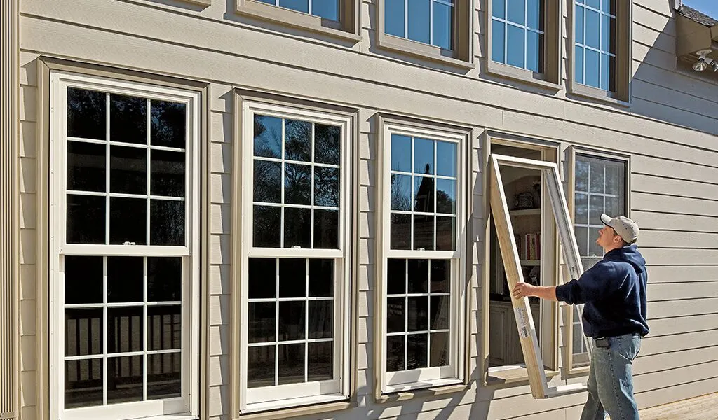 A man in a baseball cap and hoodie installs a large, white-framed window pane into a house with beige siding.