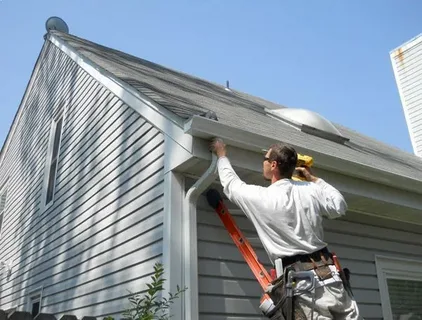 Installer on an orange ladder attaching a white aluminum gutter during a professional Seamless Gutter Installation