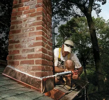Professional technician performing residential brick chimney repair and flashing sealant application on a roof